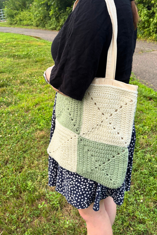 Person holding a green and white crochet tote bag outdoors on grass